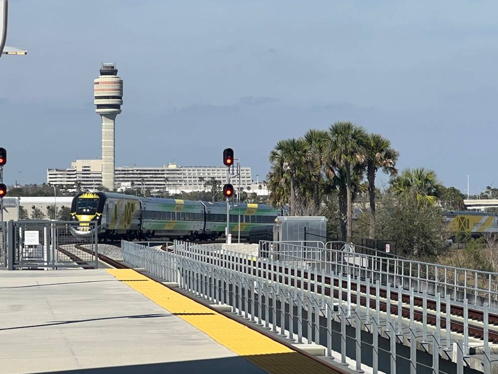 A yellow and green passenger train waits at a station platform, with a control tower, palm trees, and a nearby Black Car Service Orlando vehicle in the background.