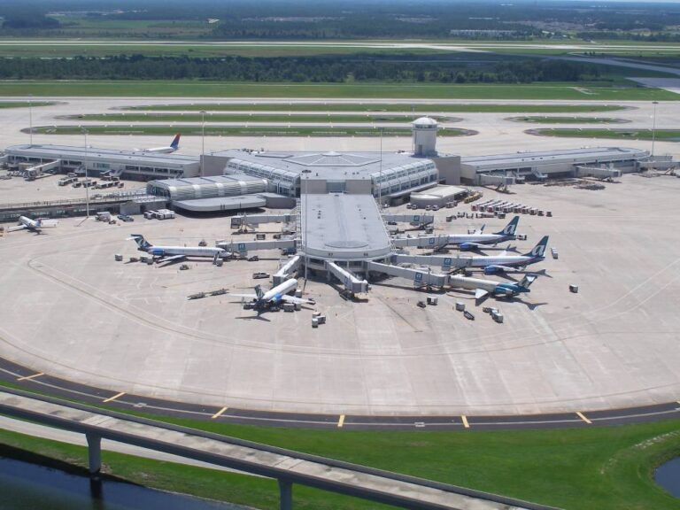 Aerial view of an airport terminal featuring Olive Luxury Ride vehicles near multiple airplanes parked at gates, with jet bridges and a control tower in the background.