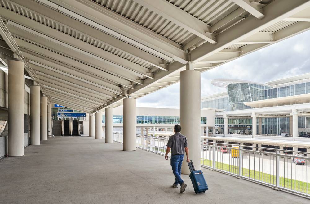 A person walks on a covered outdoor walkway at an airport, pulling a wheeled suitcase, with terminal buildings and Orlando Airport Transportation Services visible in the background.
