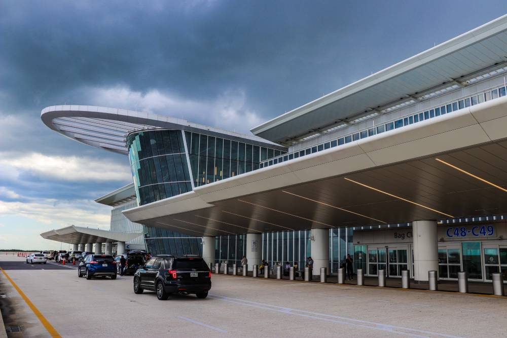 Cars are parked in front of a modern airport terminal with large glass windows and an overhanging roof under a cloudy sky, showcasing easy transportation to MCO Airport.
