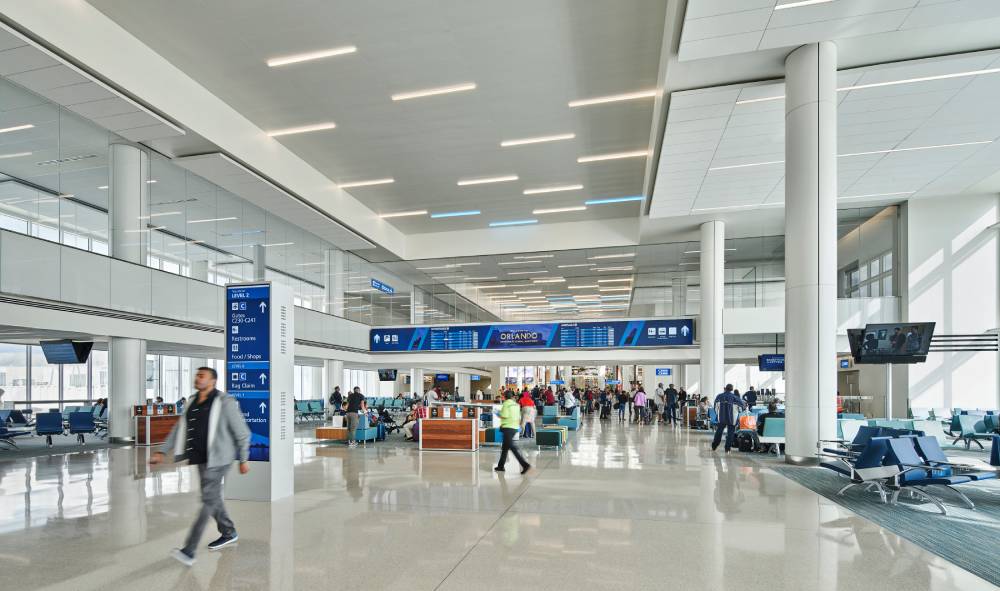 Travelers walk through a spacious, modern airport terminal with seating areas, large windows, signs, and digital flight information boards after arranging transportation to MCO Airport.