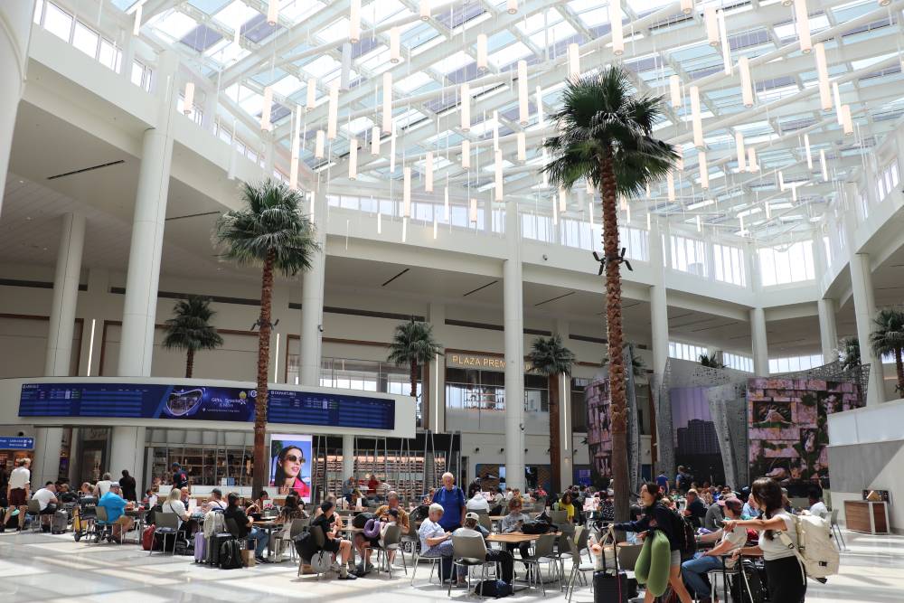 Travelers sit and walk in a spacious, sunlit airport terminal with tall glass ceilings, palm trees, digital displays, and seating areas—ideal for those awaiting Orlando Airport Transportation Services.