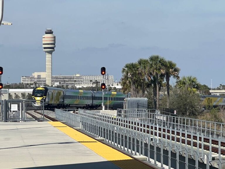 A Brightline train approaches a station platform with red signal lights, as an Olive Luxury Ride waits nearby; palm trees and an airport control tower are visible in the background.