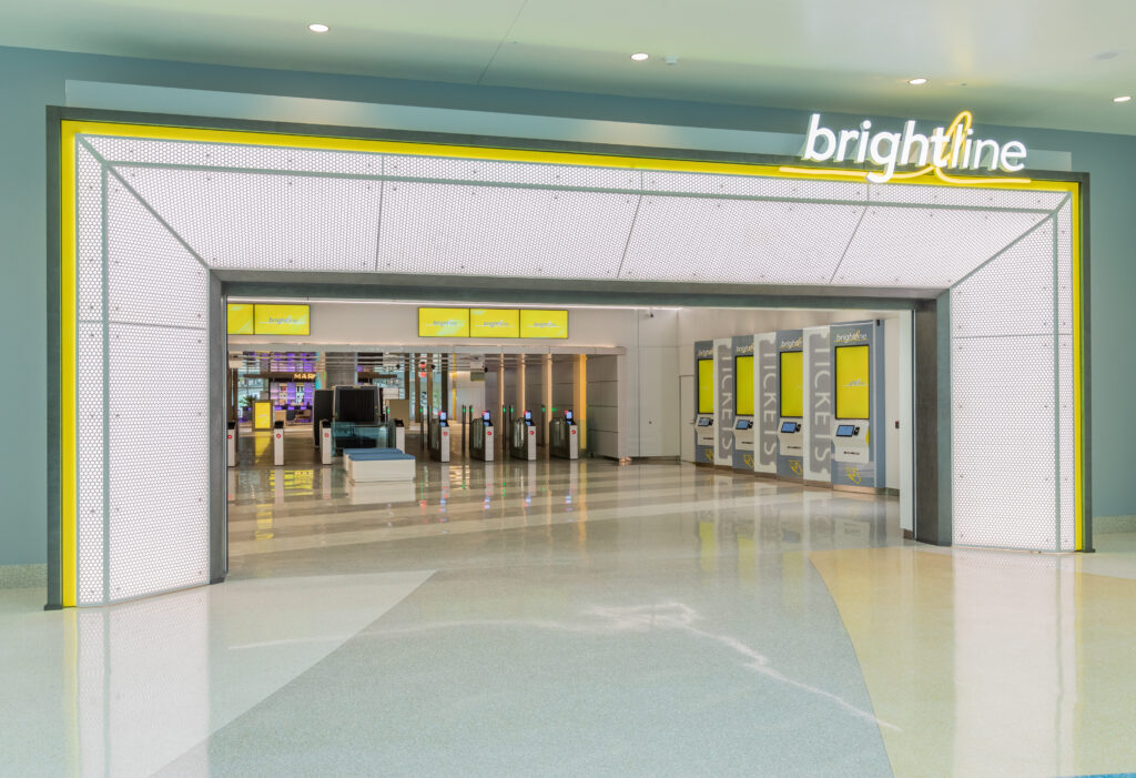 Entrance to the Brightline train station with ticket kiosks and turnstiles visible inside. The area is clean and modern, featuring yellow accent lighting and signage for transportation to MCO Airport.