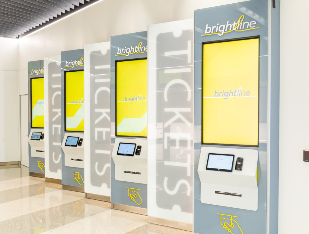 Four Brightline ticket kiosks with yellow screens and "TICKETS" labels are lined up in a clean, modern indoor setting, offering convenient transportation to MCO Airport.