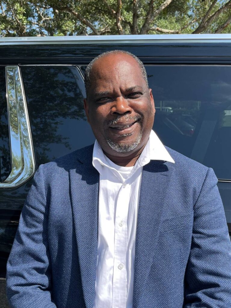 A man in a blue blazer and white shirt stands outdoors in front of a dark Car Service vehicle, smiling at the camera. Trees and sunlight are visible in the background.