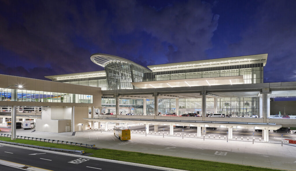 Modern airport terminal at night with glass facade, illuminated interior, parked vehicles—including those offering Car Service in Orlando Airport—and a partly cloudy sky in the background.