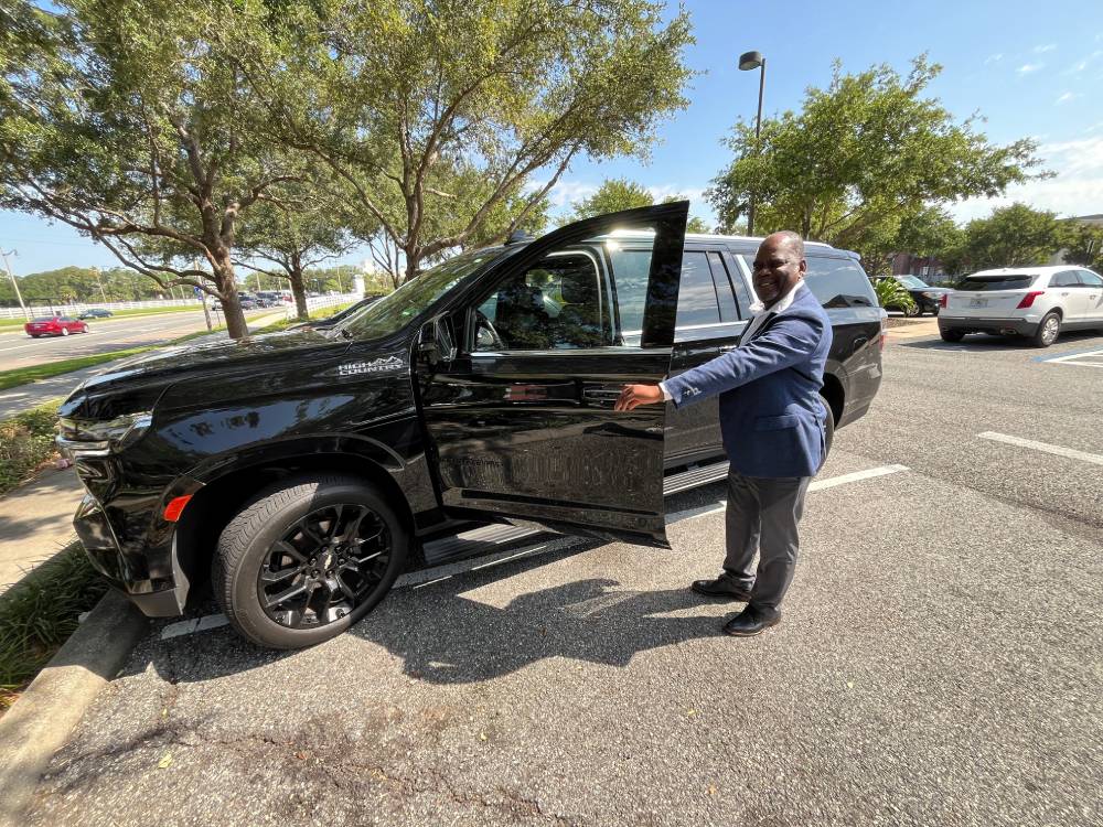 A man in a suit stands next to a black Olive Luxury Ride SUV with the driver's door open in a parking lot on a sunny day.