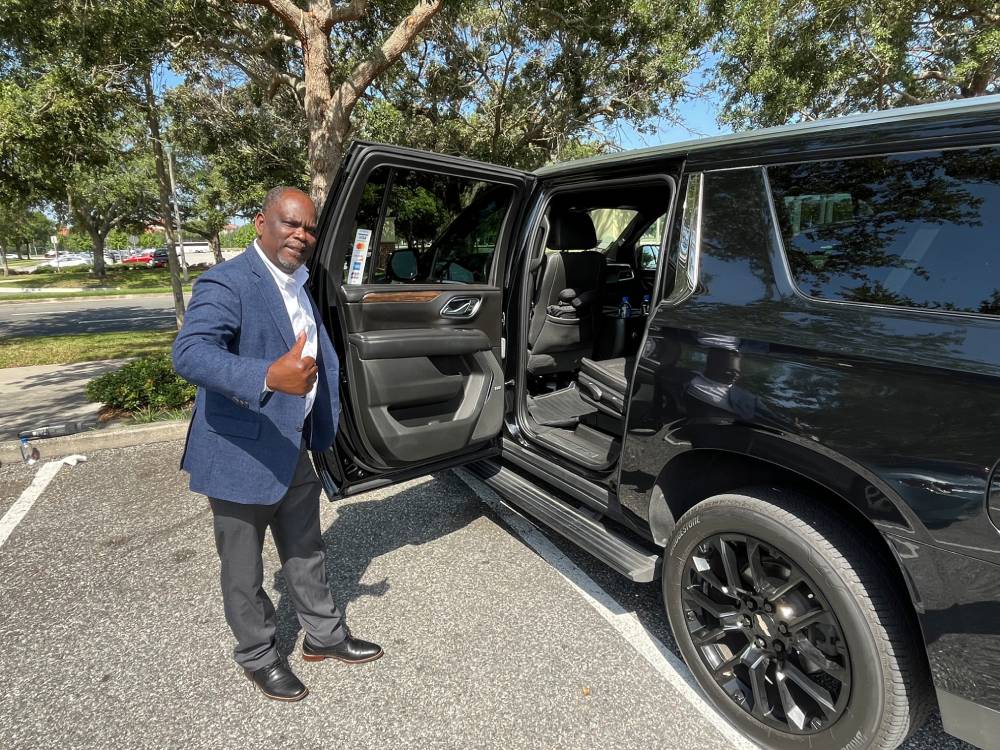 A man in a blue blazer and gray pants stands next to an open black SUV door in a parking lot, giving a thumbs-up gesture—exemplifying the professionalism of a Luxury Car Service in Orlando.