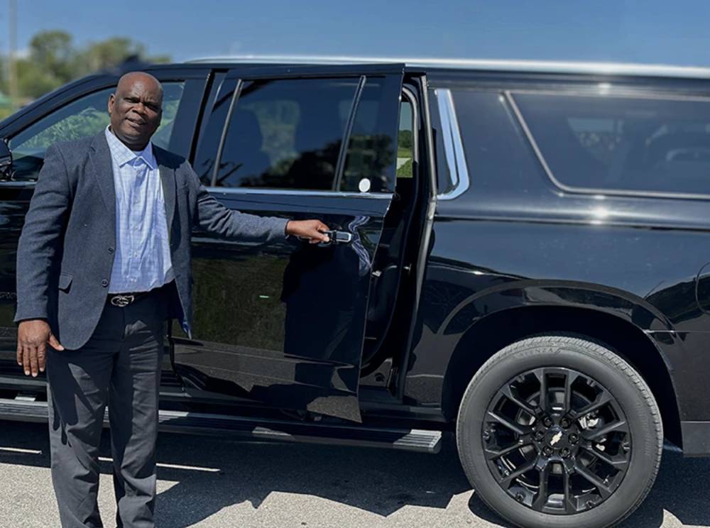 A man in a suit stands next to a black SUV with the rear door open, holding the door handle on a sunny day—exemplifying the professionalism of Car Service Orange County.