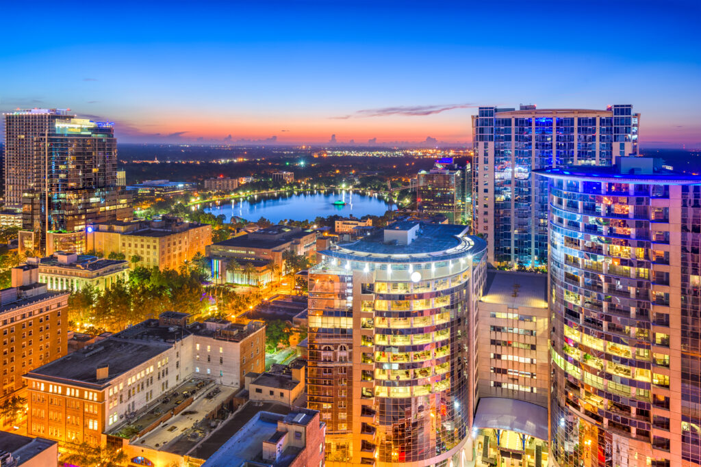 A city skyline at dusk shows high-rise buildings with lights on, streets below bustling like Car Service in Orlando Airport, and a lake surrounded by trees in the background under a blue and orange sky.