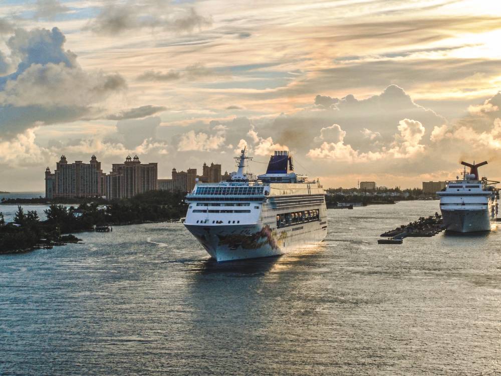 A large cruise ship sails towards port with another ship docked nearby under a partly cloudy sunset sky, as resort buildings and transportation from MCO to Port Canaveral complete the lively scene in the background.