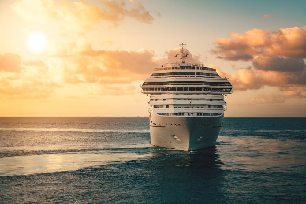 A large cruise ship sails on calm ocean water at sunset, with clouds in the sky and the sun low on the horizon—marking the perfect end to a journey that began with transportation from MCO to Port Canaveral.