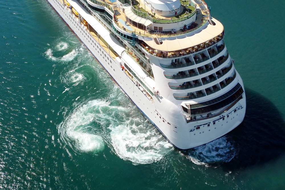A large white cruise ship is turning in blue-green water, creating visible swirling wake patterns behind its stern—much like the journey travelers take with transportation from MCO to Port Canaveral before setting sail.