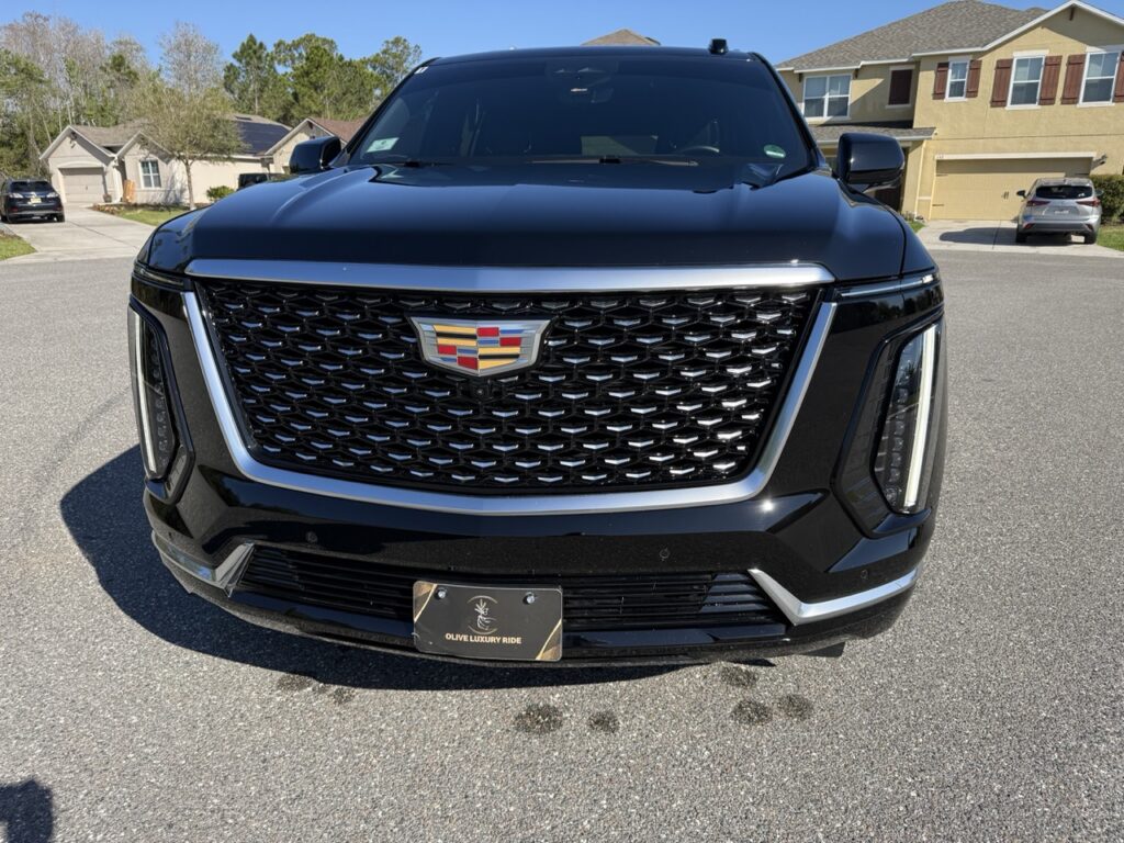 Front view of a black Cadillac SUV parked on a residential street, showing the grille, emblem, and license plate.